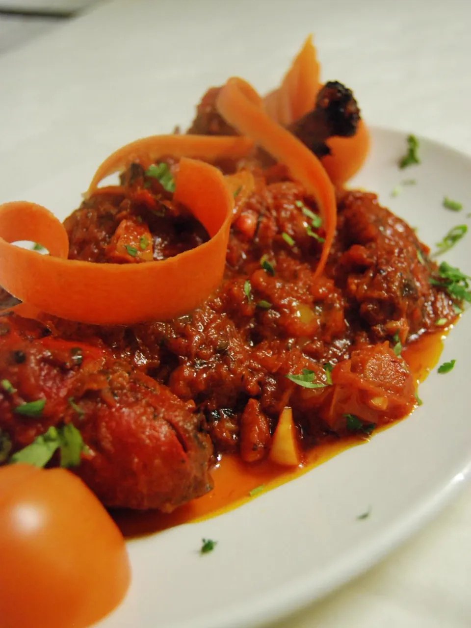 A white plate of meat in a thick red sauce, with a curl of carrot and half a tomato.
